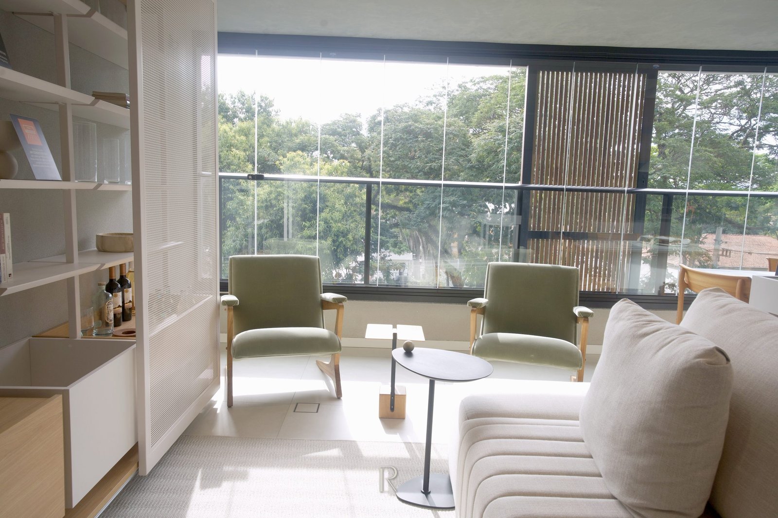 Estar do apartamento decorado, com poltronas junto à face de vidro e as árvores da rua